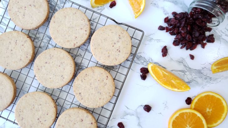 Cranberry orange shortbread sugar cookies on a cooling rack, next to dried cranberries and orange slices. 