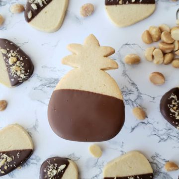 a single pineapple shaped, macadamia nut cookie, dipped in chocolate, surrounded by macadamia nuts and smaller cookies