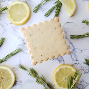 A single lemon rosemary cookie surrounded by lemon slices and sprigs of fresh rosemary