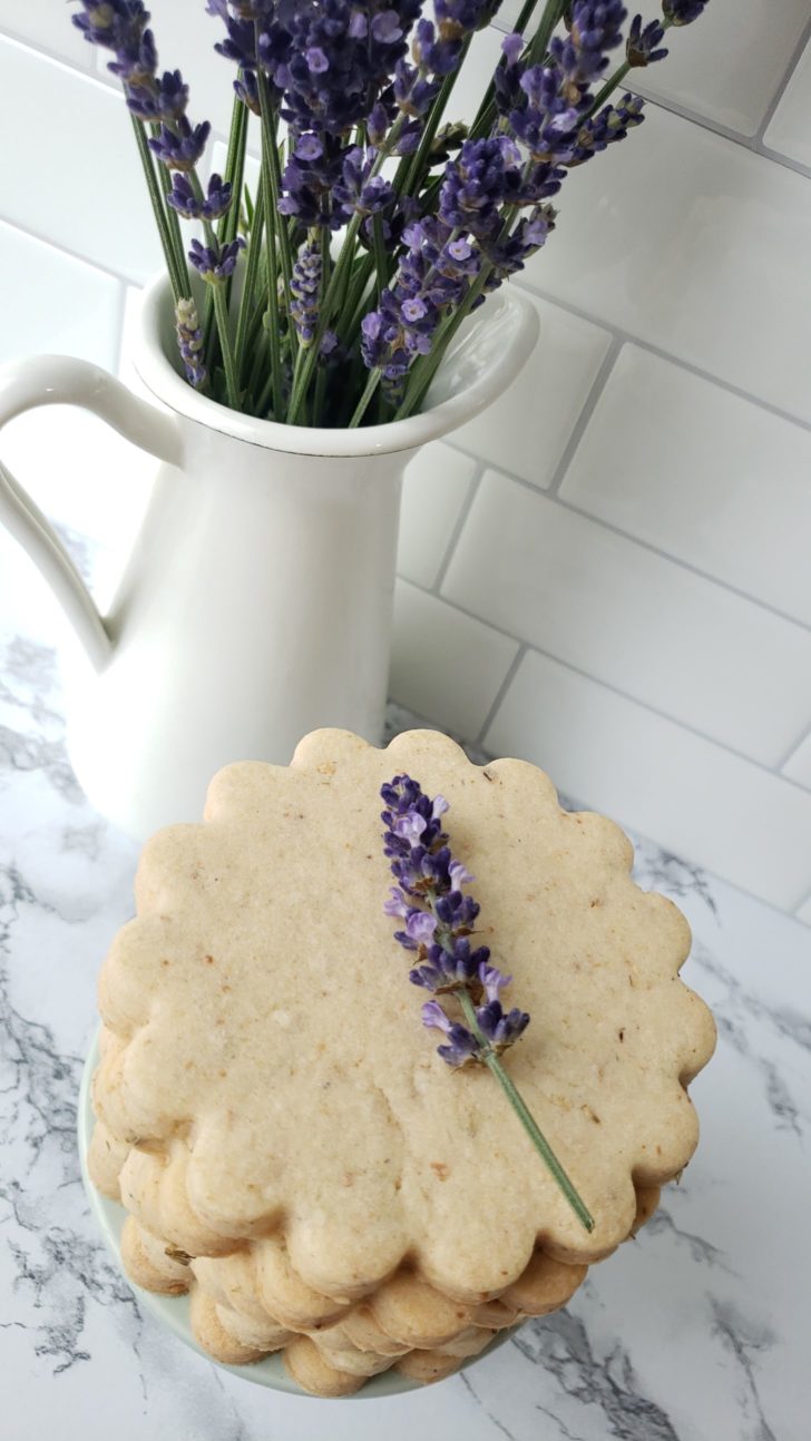 Lavender shortbread sugar cookies stacked in front of a vase with lavender flowers. 