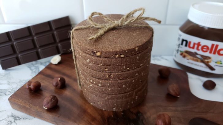 Stacked chocolate hazelnut cookies tied with twine, surrounded by hazelnuts, bar of chocolate, and a jar of nutella. 