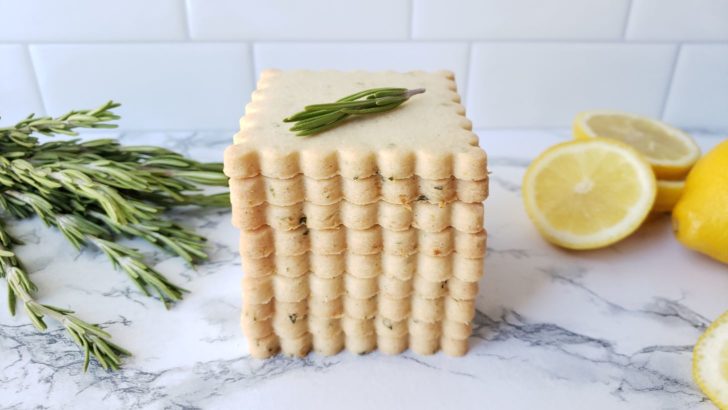 Lemon rosemary shortbread sugar cookies stacked in front of fresh sliced lemon and a bundle of fresh rosemary. 