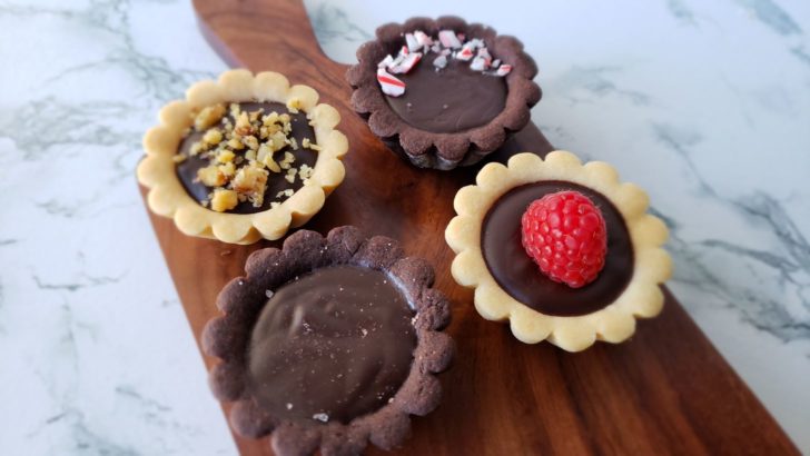 4 mini ganache tarts sitting on a small wooden cutting board on a marble surface. The tarts are topped with sea salt, candy cane crumbles, chopped walnuts or a single raspberry. 