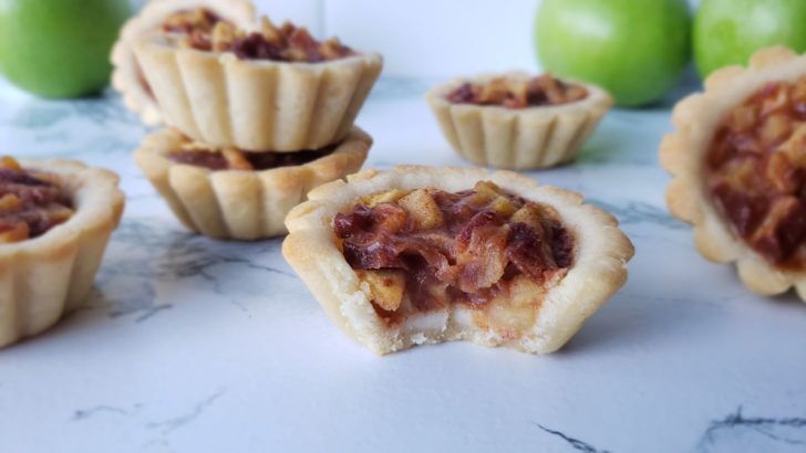 An apple pie tartlet with a bite in the forefront, surrounded by more apple pie tartlets. 
