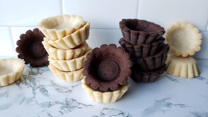 Empty chocolate and vanilla shortbread tartlet shells stacked on a marble counter with a white subway tile backsplash in the background. 