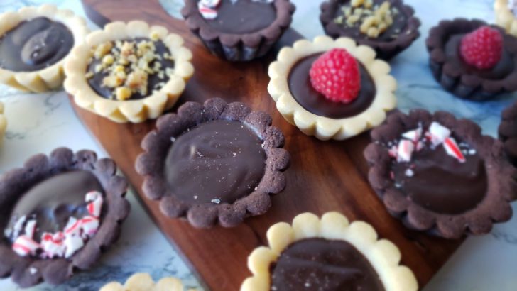Mini ganache tarts sitting on a small wooden cutting board on a marble surface. The tarts are topped with sea salt, candy cane crumbles, chopped walnuts or a single raspberry. 