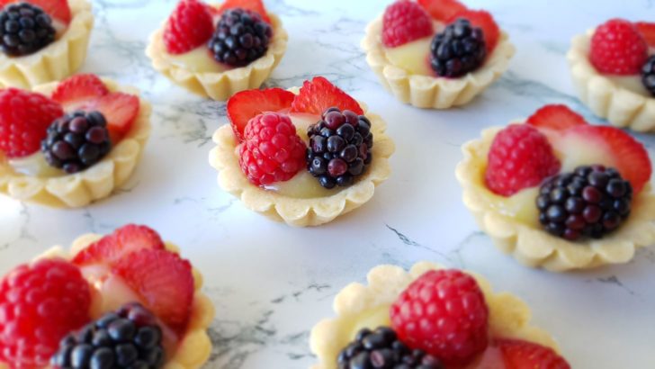 Fruit tarts with lemon curd, blackberries, raspberries, and strawberries, sitting on a marble surface. 