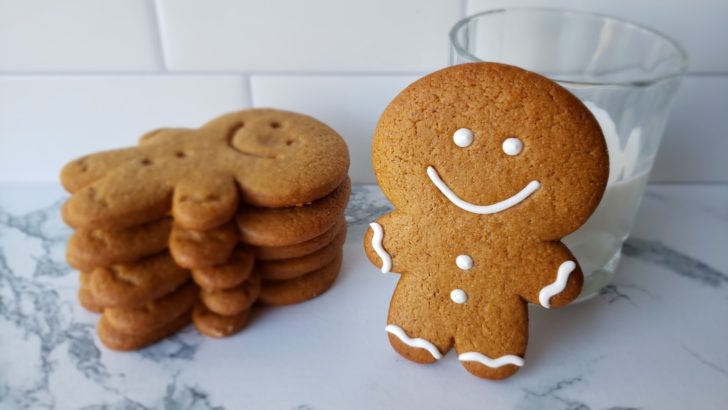 A single gingerbread man cookie leaning against a glass of milk next to a stack of gingerbread men cookies. 