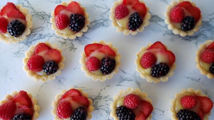 Fruit tarts with lemon curd, blackberries, raspberries, and strawberries, sitting on a marble surface. 