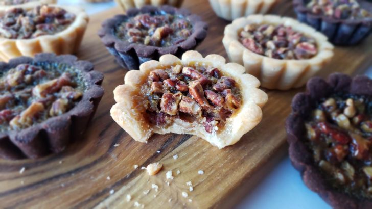 Mini pecan pie tarlets on a wooden cutting board. One of the tartlets has a bite in it. 