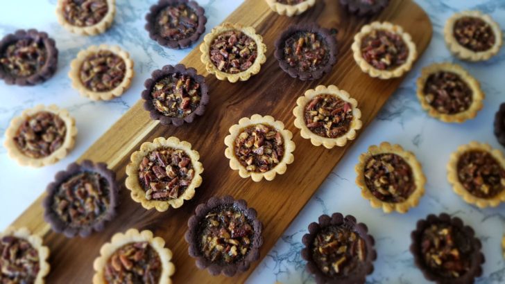 Mini pecan pie tarlets on a wooden cutting board on a marble surface. 