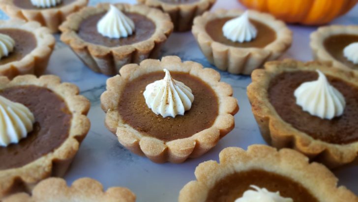 Close up of mini pumpkin pie tarts topped with whipped cream sitting on a marble surface.