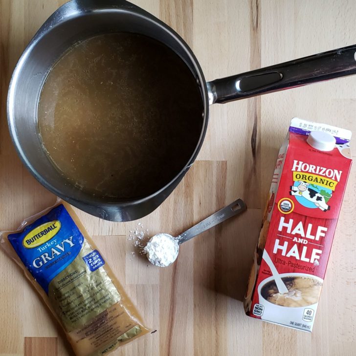 Ingredients for turkey gravy sitting on a wooden butcher block. A saucepan roast turkey drippings, a packet of gravy from a frozen turkey, a tablespoon of cornstarch, and a quart container of half-and-half. 