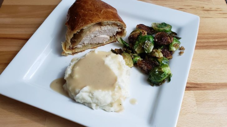 A plate with a slice of turkey wellington, mashed potatoes and gravy, and sauteed brussels sprouts. 