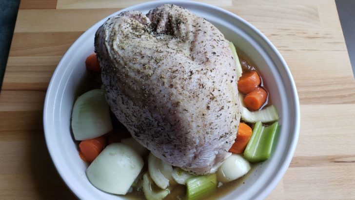 A large pie pan with a raw turkey sitting on top of vegetables and turkey stock. 