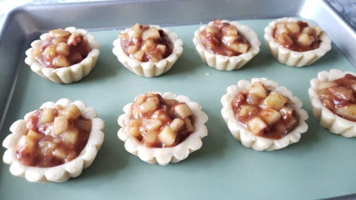 Pre-baked apple pie tartlets sitting on a cookie sheet. 